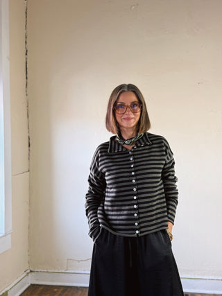 Woman in olive and black striped button up cardigan and olive silk scarf and black pants in front of cream wall.
