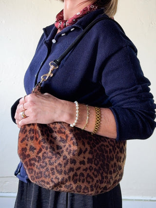 A person wearing a navy cardigan and patterned scarf carries a leopard-print shoulder bag with a black strap and gold-tone hardware against a plain off-white wall.