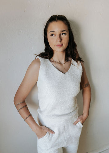 Woman wearing a white sleeveless top and pants against a plain wall.