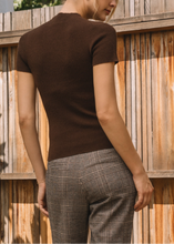 Load image into Gallery viewer, Person wearing a brown top and plaid pants standing against a wooden fence.
