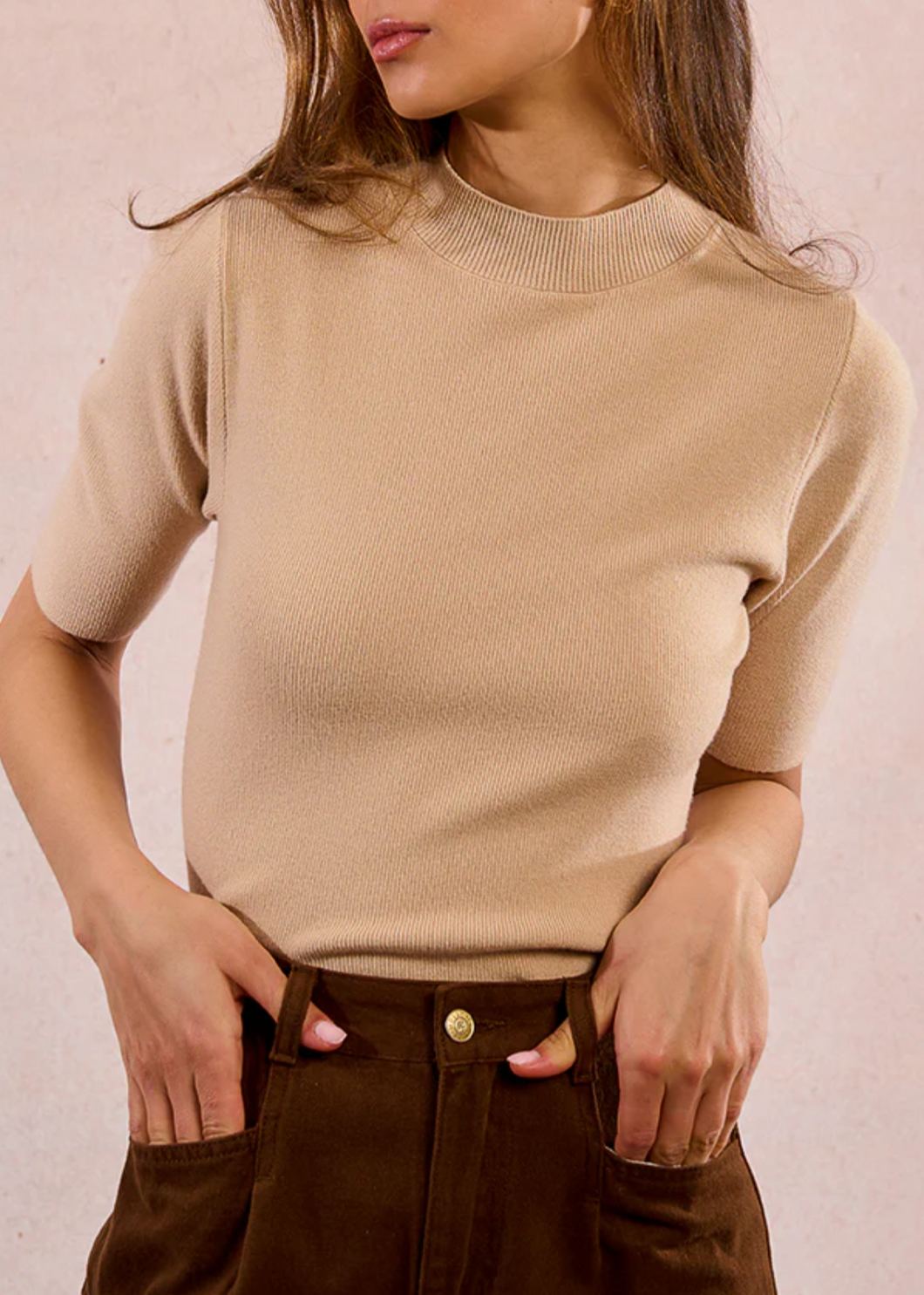 Woman wearing a beige sweater and brown pants against a plain background